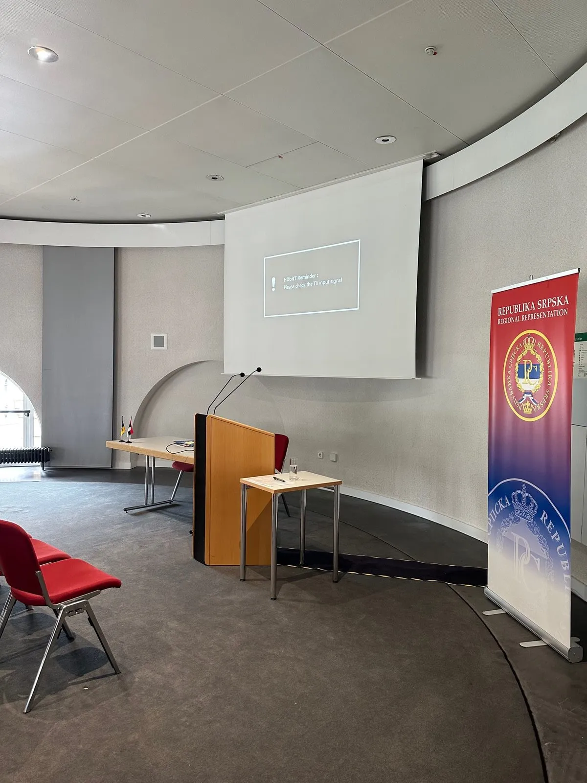 Der Konferenzraum vor Beginn: Rednerpult mit Mikrofon, daneben ein Tisch mit deutschen Tischfähnchen, an der Wand eine Leinwand. Rechts steht ein rot-blauer Aufsteller mit dem Wappen der Republika Srpska und der Aufschrift „Republika Srpska — Regional Representation".