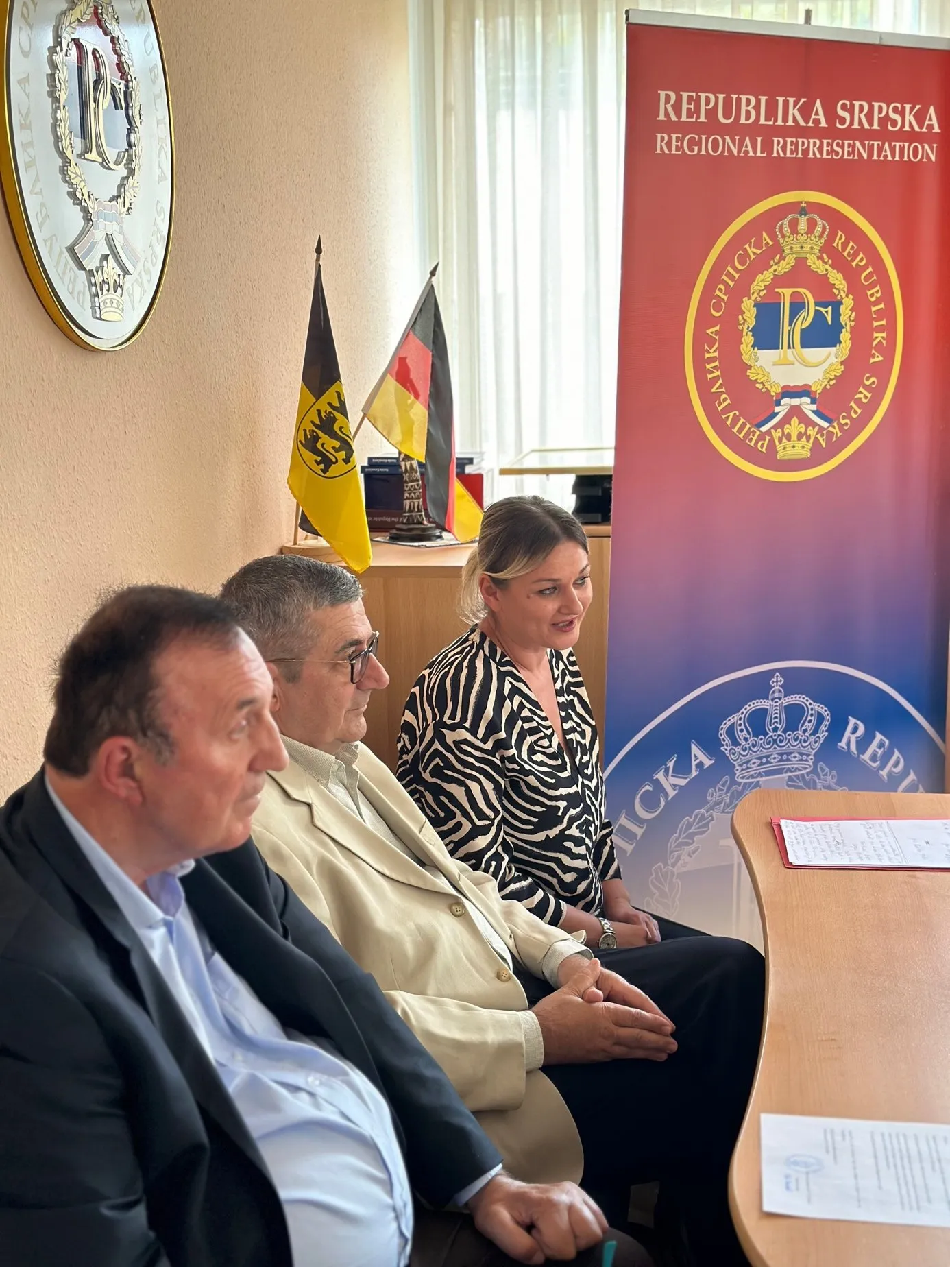 Drei Gäste sitzen vor einem roten Aufsteller mit der Aufschrift Republika Srpska Regional Representation, daneben die Flaggen Baden-Württembergs und Deutschlands.
