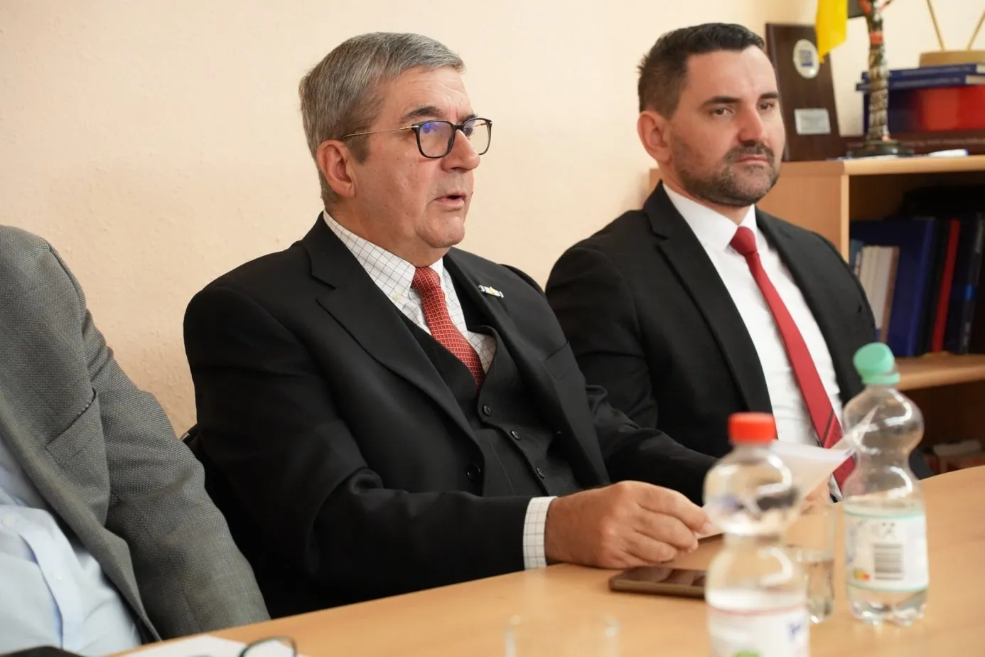 Zwei Herren in dunklen Anzügen mit roten Krawatten sitzen nebeneinander am Konferenztisch, vor ihnen zwei Wasserflaschen, im Hintergrund ein Aktenregal.