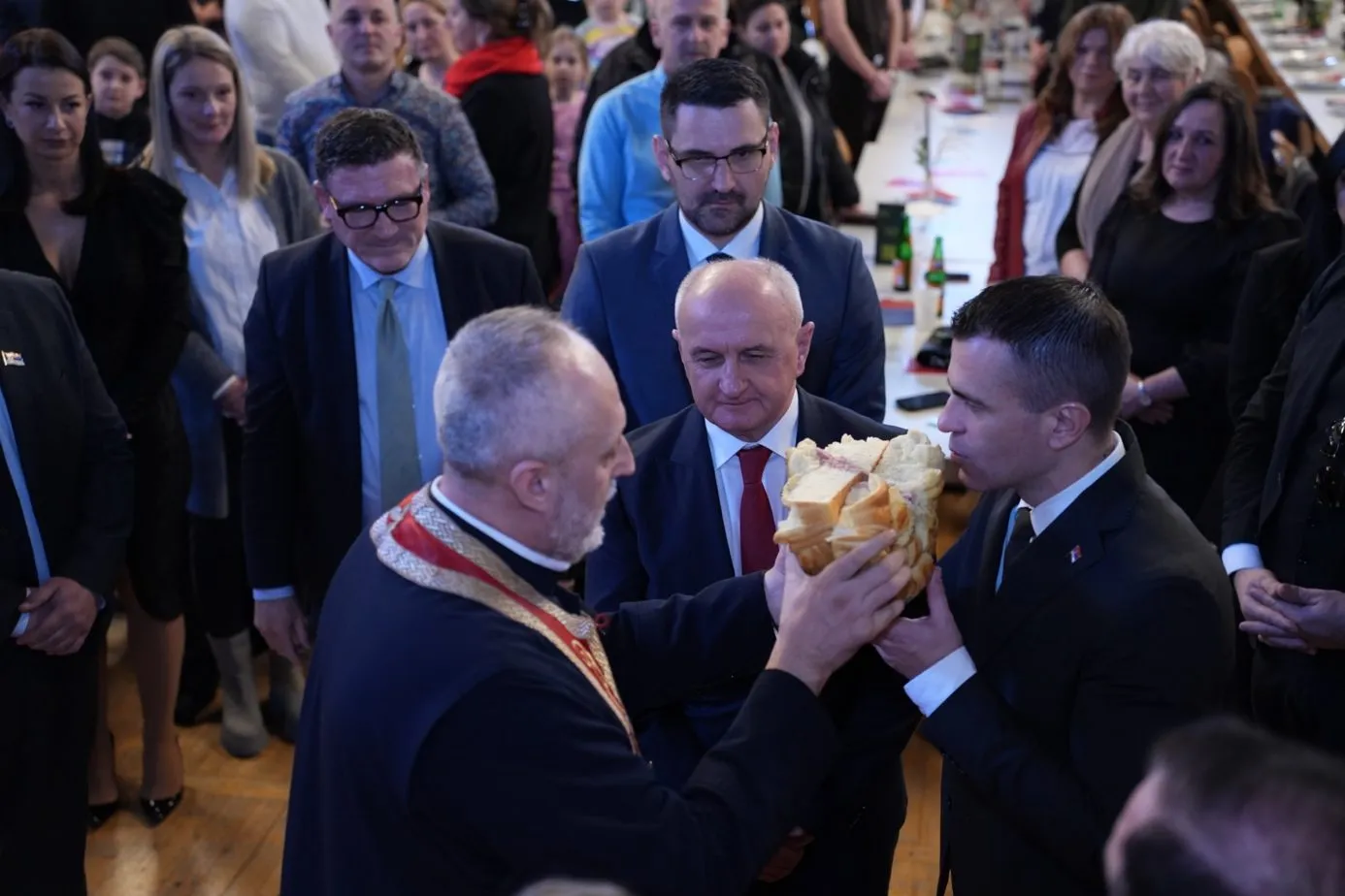 Ein orthodoxer Geistlicher in Stola bricht das Festbrot gemeinsam mit zwei Männern auseinander, umringt von stehenden Gästen.