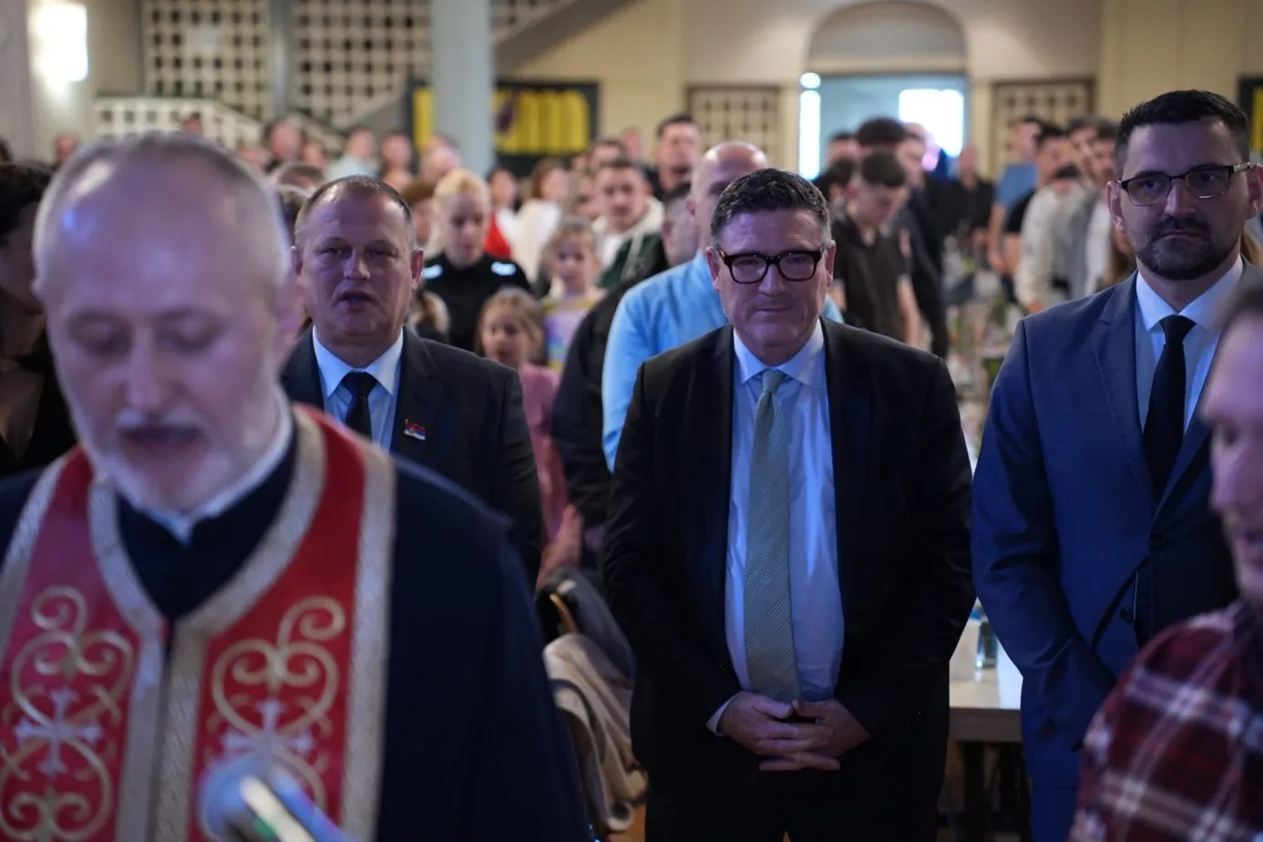 Gäste stehen während der Zeremonie in der Halle. Im unscharfen Vordergrund der Geistliche mit rot-weiß besticktem Gewand.