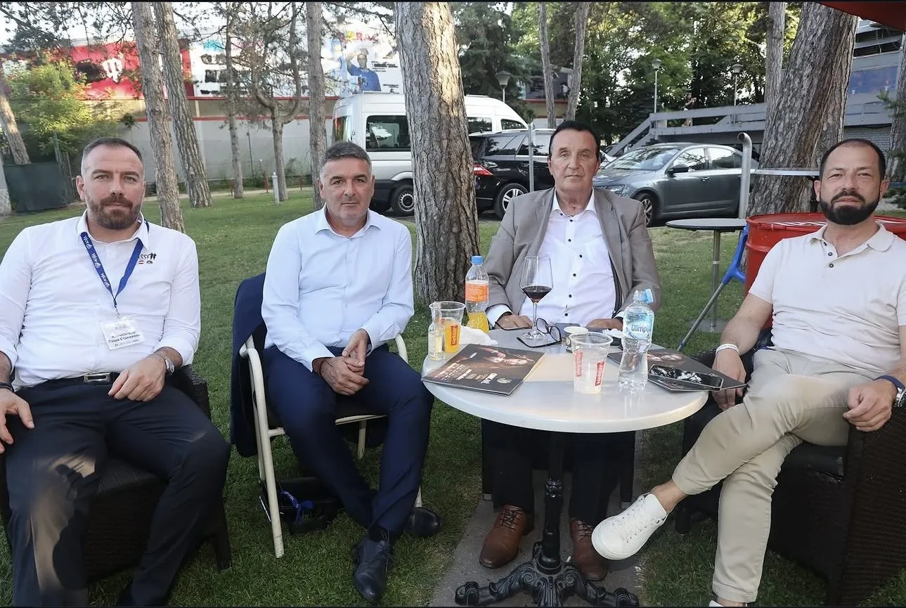 Vier Männer sitzen im Freien zwischen Kiefern an einem runden Gartentisch, auf dem Programmhefte und Getränke stehen.