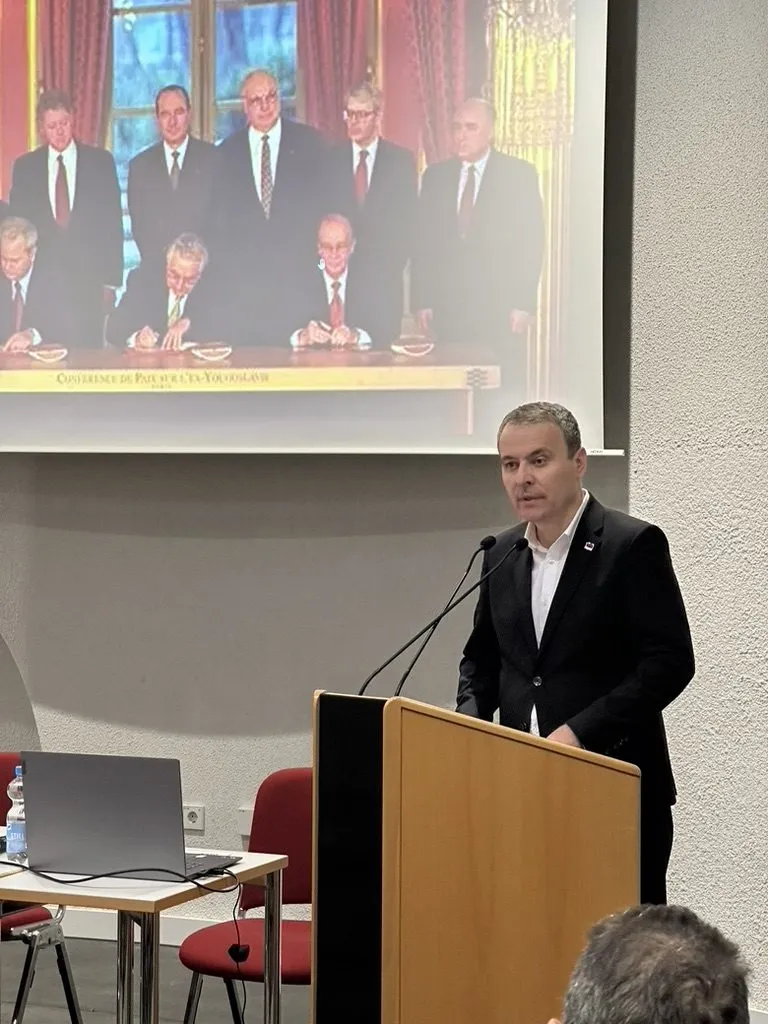 Ein Redner steht an einem Holzpult mit zwei Mikrofonen. Hinter ihm die Projektion der Unterzeichnungszeremonie.