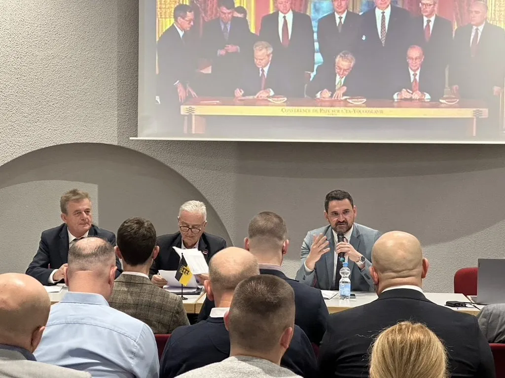 Drei Teilnehmer am Podiumstisch, der rechte spricht in ein Handmikrofon. Im Vordergrund die Hinterköpfe der Zuhörer, darüber die Projektion.