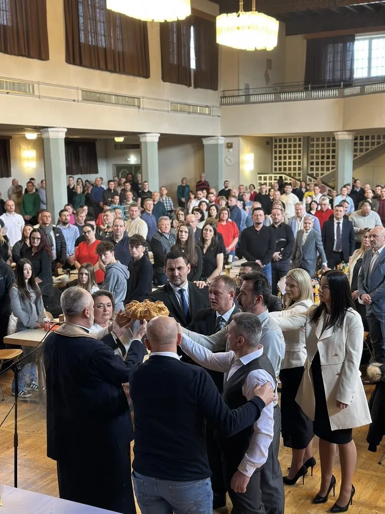 Mehrere Gäste halten das runde Festbrot gemeinsam in die Höhe und drehen es, während der Geistliche das Brot segnet. Ringsum steht die Festgesellschaft dicht gedrängt.