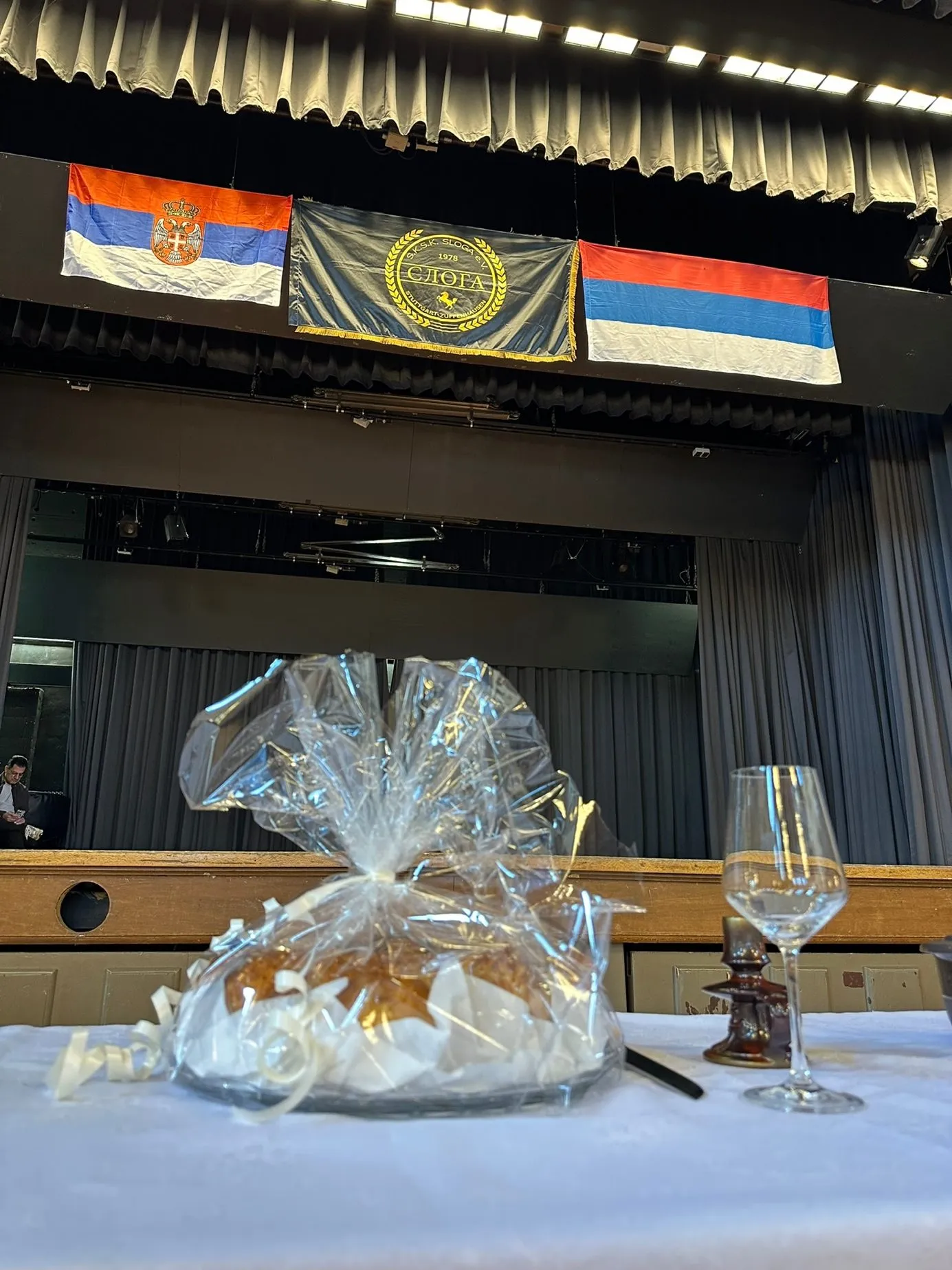 Vor der Bühne hängen nebeneinander die Flagge der Republik Serbien, die Vereinsfahne des S.K.S.K. Sloga Stuttgart-Zuffenhausen von 1978 und die Flagge der Republika Srpska. Im Vordergrund auf weißem Tischtuch ein in Klarsichtfolie eingeschlagenes Festbrot und ein Weinglas.