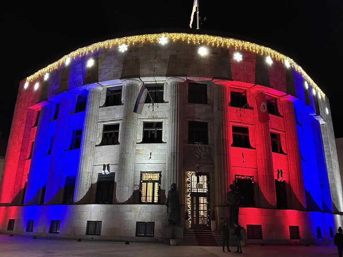 Ein repräsentatives Verwaltungsgebäude bei Nacht, angestrahlt in Rot, Blau und Weiß, auf dem Dach weht eine Flagge.