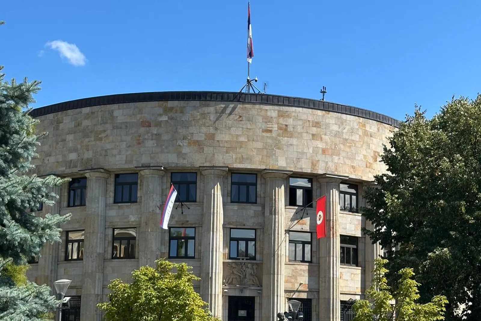 Das Gebäude der Regierung der Republika Srpska in Banja Luka bei Tag, eine helle Steinfassade mit Säulen und gerundeter Front, davor Bäume und ein Platz, auf dem Dach die Flagge der Republika Srpska.